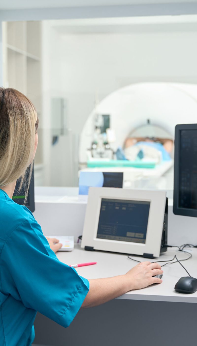 Long-haired woman in special clothes sits at a table in the tomography room and directs the procedure.